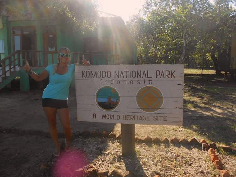 Blonde girl with sunglasses and blue top doing thumbs up by Komodo National Park Indonesia sign